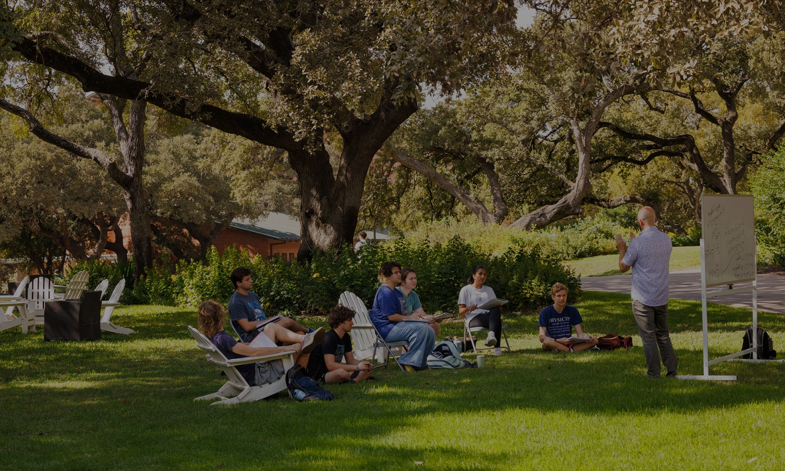 students sit in chairs outside under a tree listening to a mathematics lecture