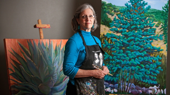 Polly Jackson Spencer, in a paint-stained apron, stands in front of two bright-colored tree paintings