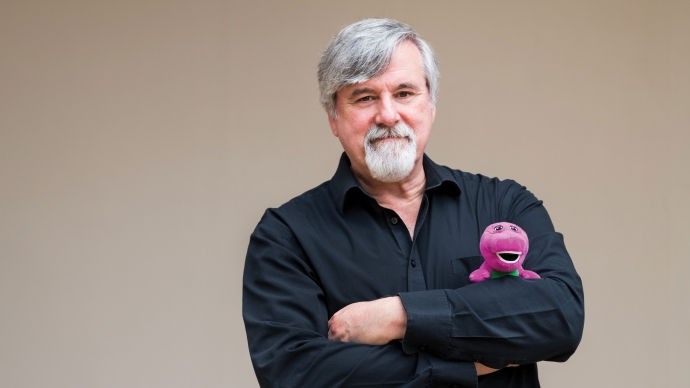 bob west in black shirt standing in front of a tan background with his arms crossed, holding a barney plush doll
