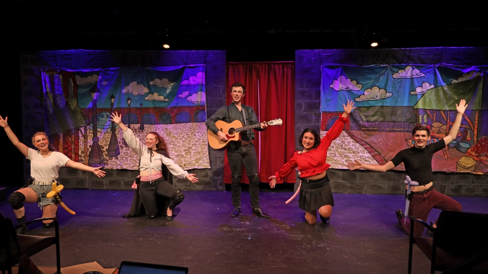 five performers on stage, one in the center holding a guitar, dance to a musical number in the Attic Theater