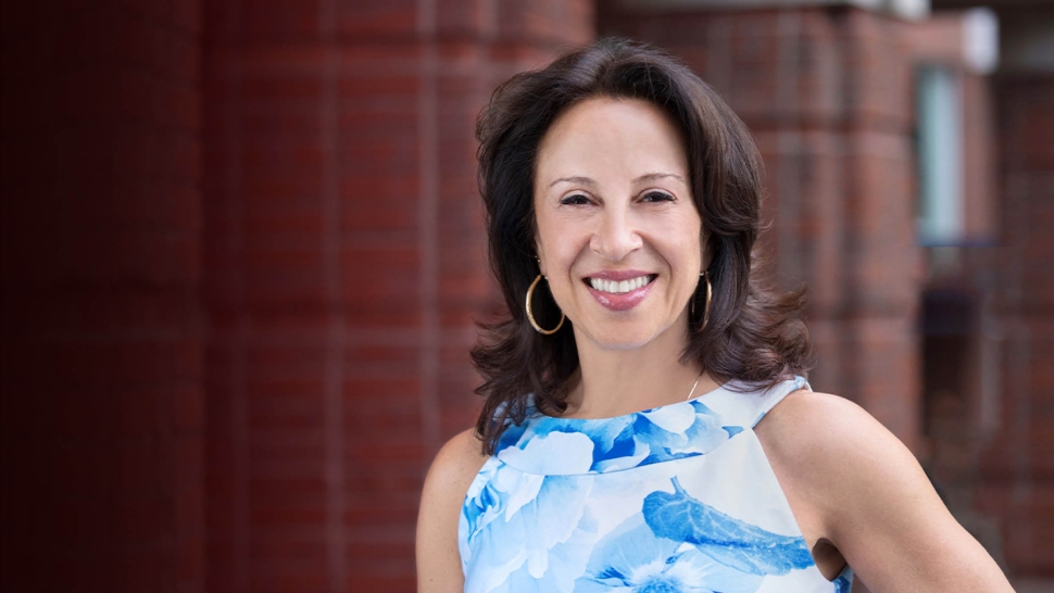 Headshot of Maria Hinojosa smiling in front of a dark red background.