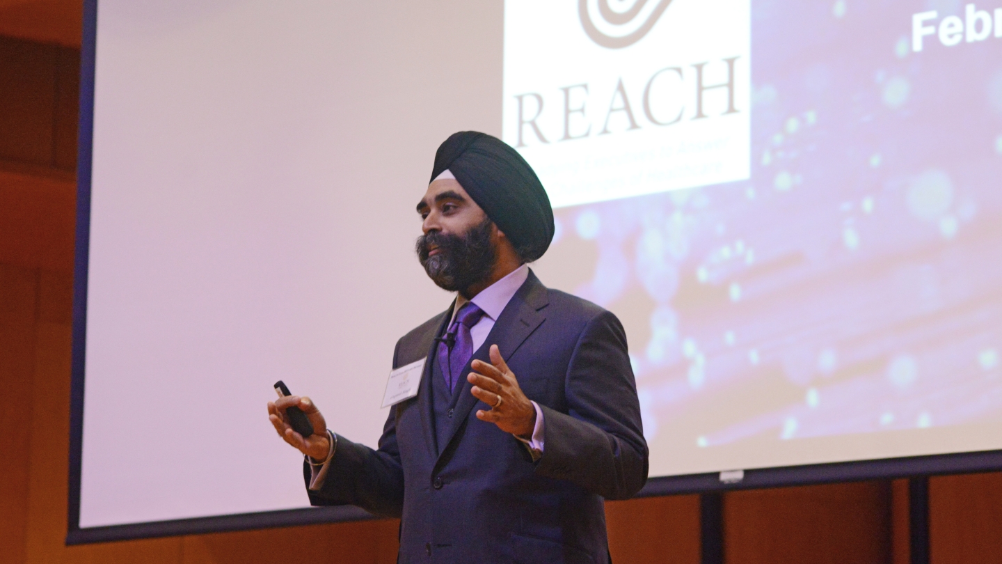 a man gives a presentation in front of a screen with the REACH Symposium logo behind him