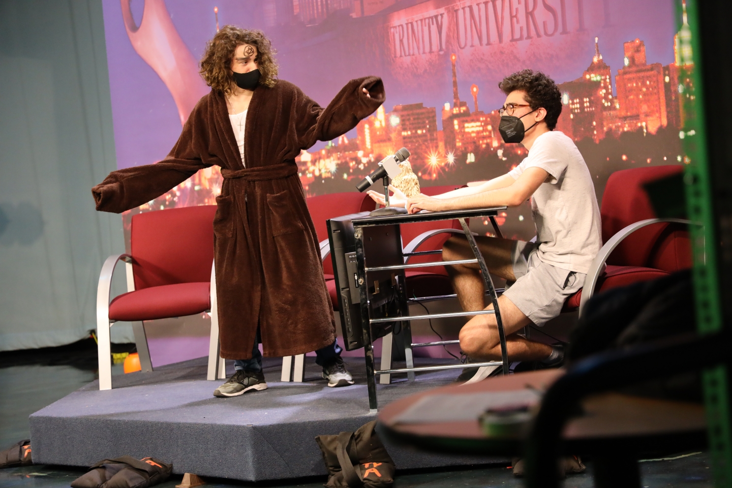 Two students on TigerTV set. Student on right sitting at a makeshift desk while student on left is wearing a robe and standing