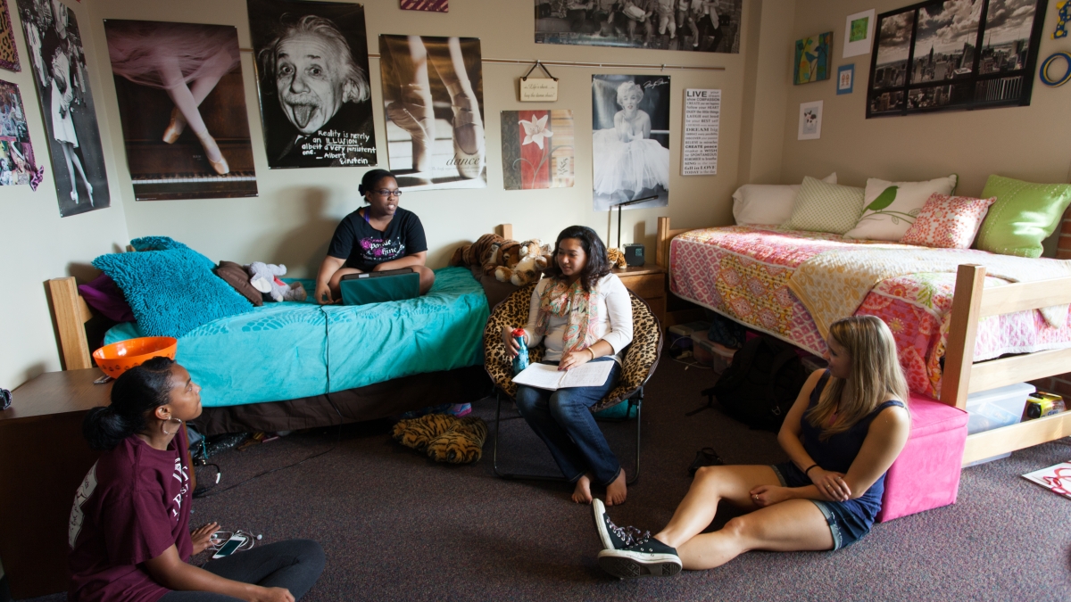 Four students, one sitting on a bed and the other three sitting on the floor in their dorm room having a conversations
