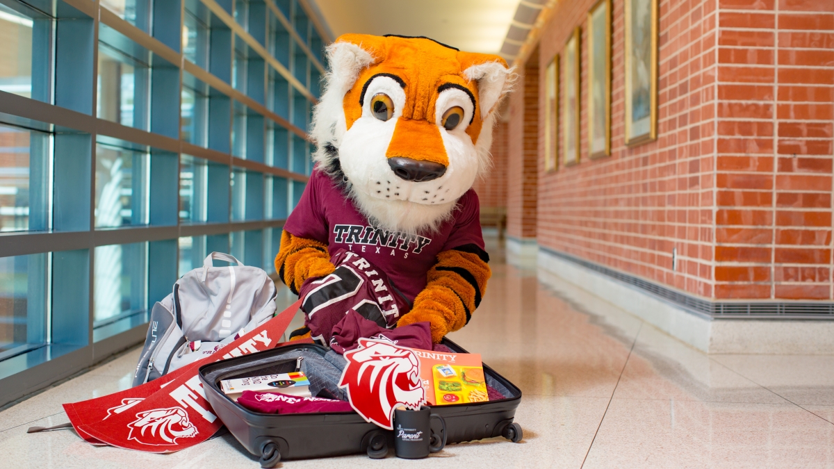 the LeeRoy Tiger mascot poses with an open suitcase full of Trinity swag